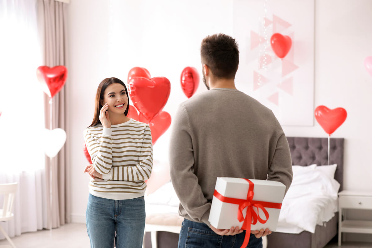 Homem segurando caixa surpresa nas costas para presentear mulher. O quarto está decorado de balões vermelhos.
