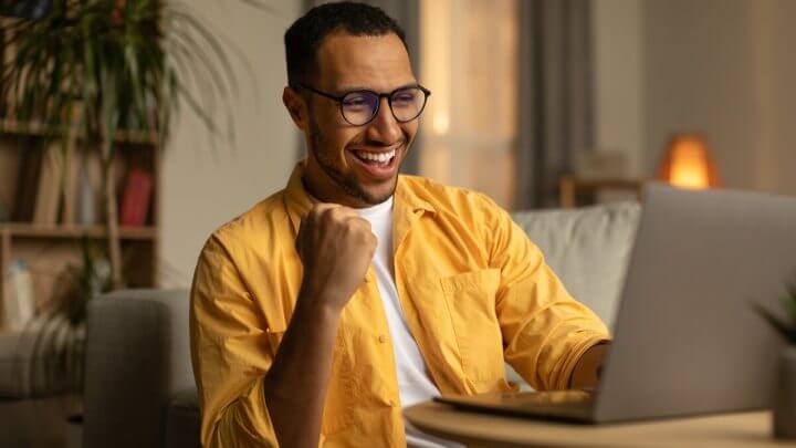Homem com blusa branca e amarela sentado em frente a um computador sorrindo