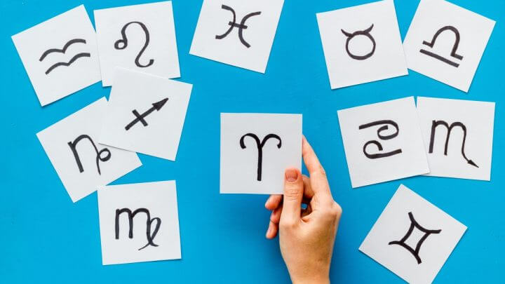Mão feminina segurando um papel com o signo de Áries em um fundo azul com desenhos dos doze signos do zodíaco