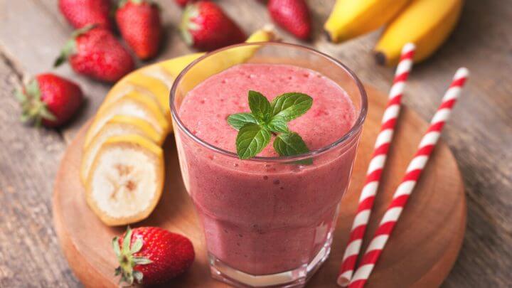 Copo com vitamina rosa decorado com folhas de hortelã em cima de uma tábua de madeira com morangos e rodelas de banana ao seu redor