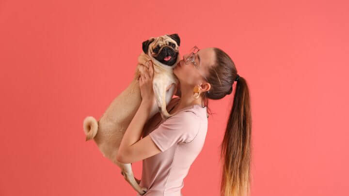 Mulher segurando um cachorro e o beijando em frente a um fundo rosa