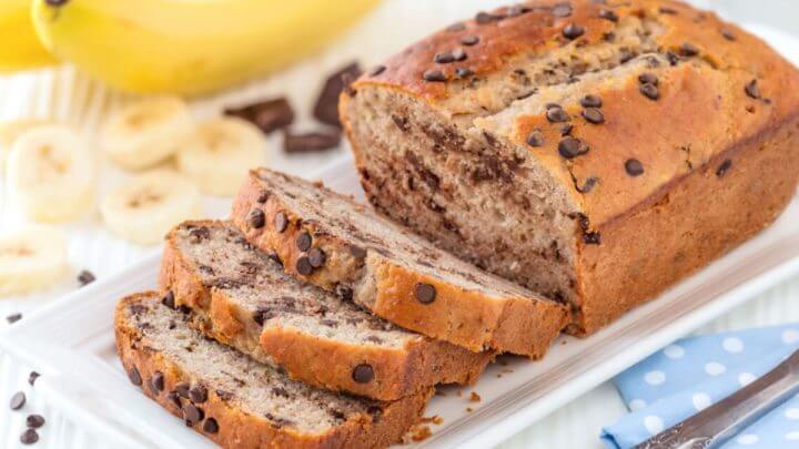 Pão de banana caseiro com lascas de chocolate fatiado em uma bandeja branca com ingredientes ao fundo