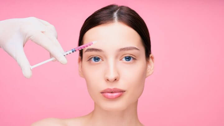 Mulher em fundo rosa aplicando botox na testa