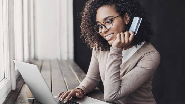 Mulher usando óculos sentada segurando um cartão e mexendo em um notebook