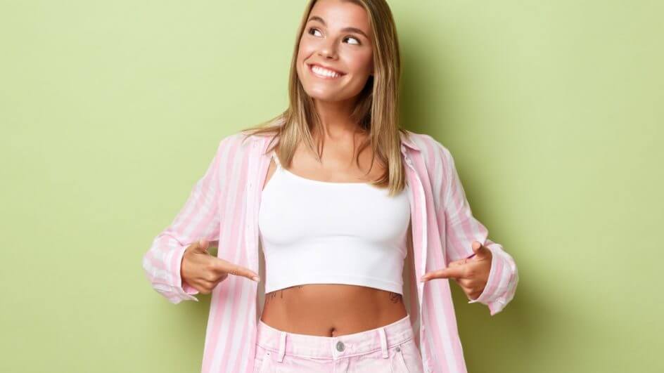 Mulher vestindo blusa branca , short e casaco rosa sorrindo e apontando para a barriga