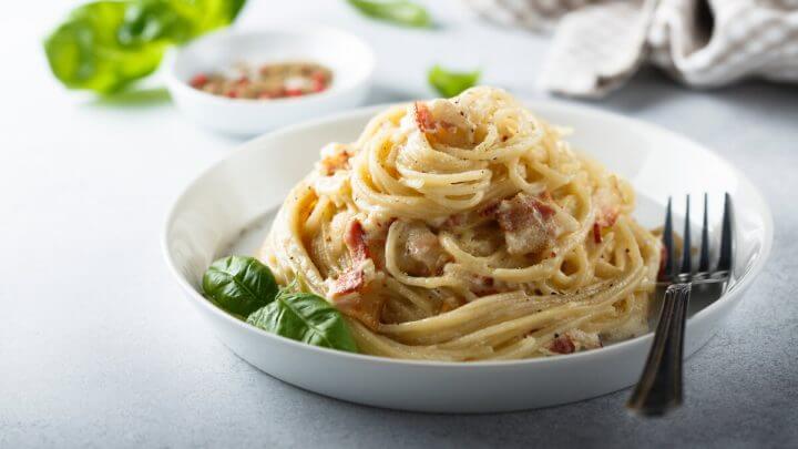 Prato branco com macarrão à Carbonara decorado com folhas em cima de uma mesa branca
