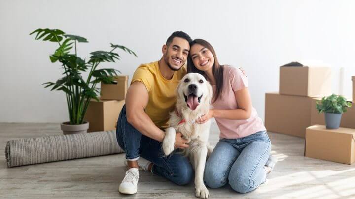 Homem e mulher abraçados com um cachorro em uma sala de estar com caixas de papelão