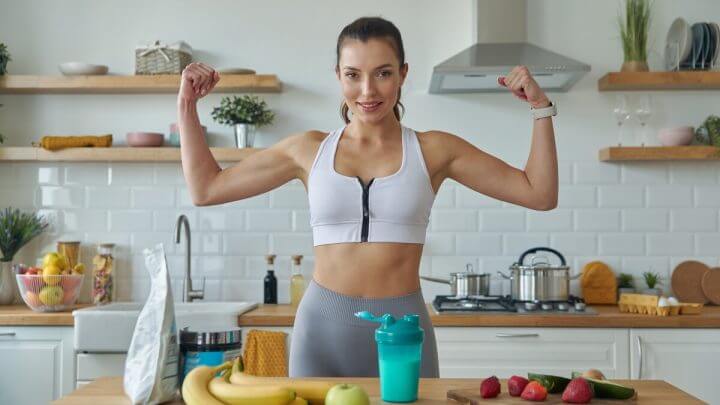 Mulher mostrando os músculos do braço em uma cozinha com mesa com frutas e whey protein