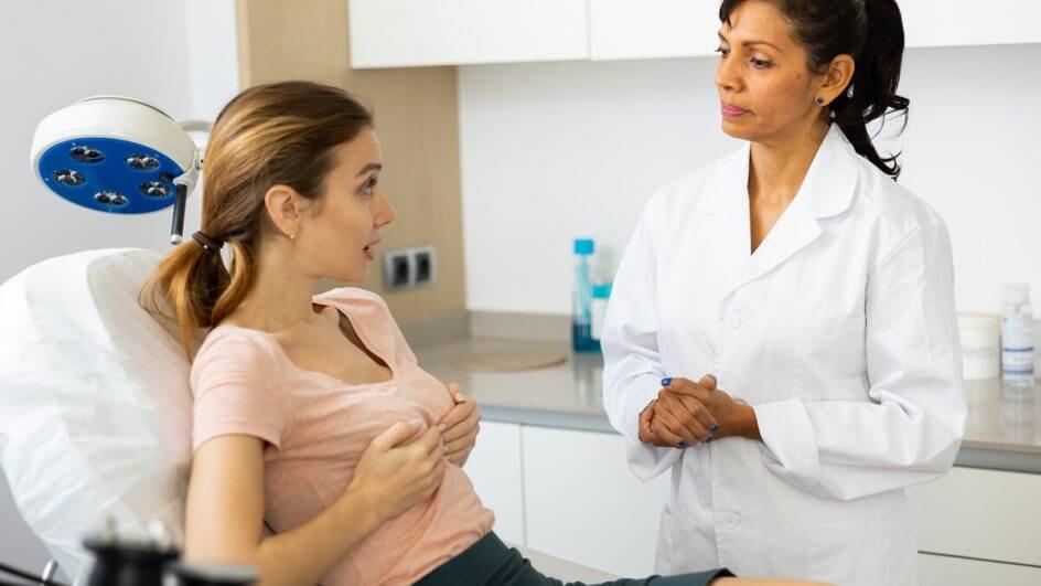 Mulher deitada com as mãos no seio conversando com uma médica