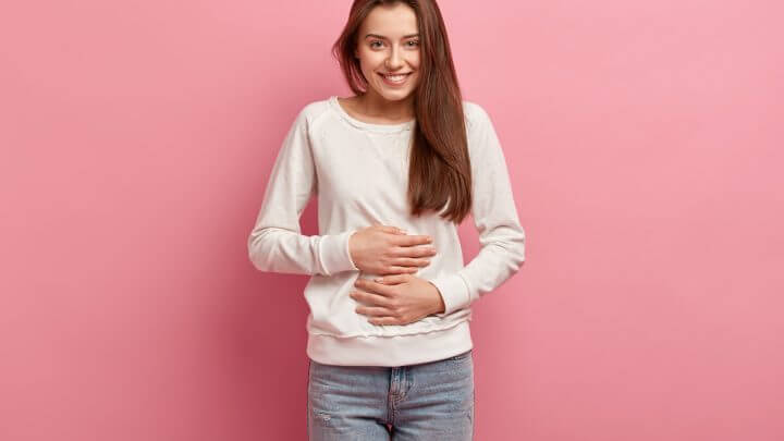 Mulher vestindo blusa branca com as mãos na barriga em frentea um fundo rosa