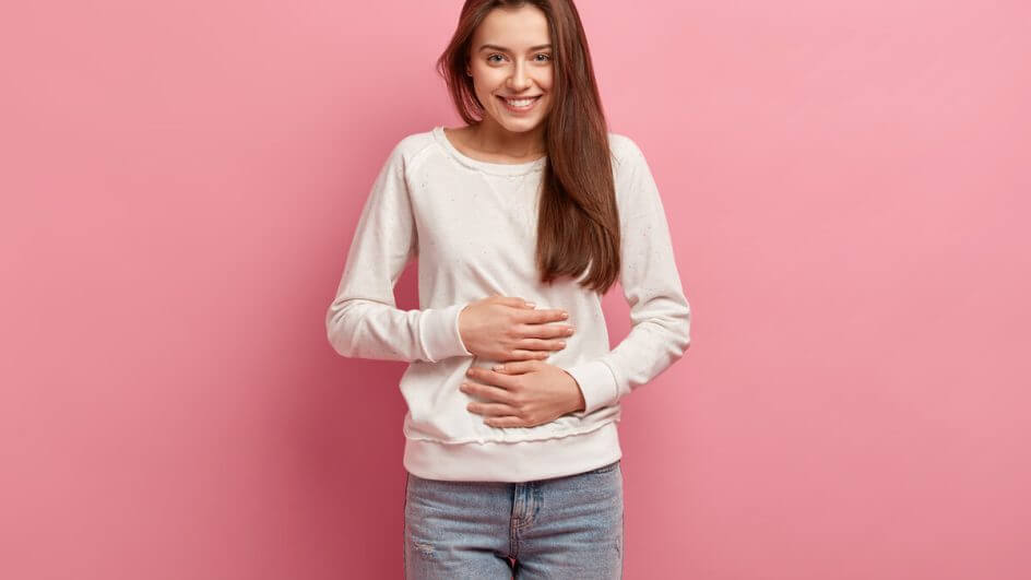 Mulher vestindo blusa branca com as mãos na barriga em frentea um fundo rosa