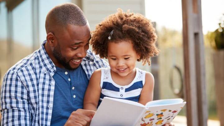 Pai lendo livro infantil com filha. Os dois estão sorrindo