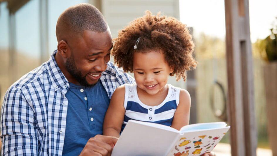 Pai lendo livro infantil com filha. Os dois estão sorrindo