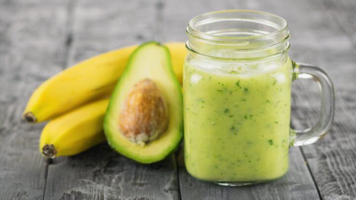 Copo de vidro com vitamina de abacate em cima de uma mesa de madeira com duas bananas e um abacate
