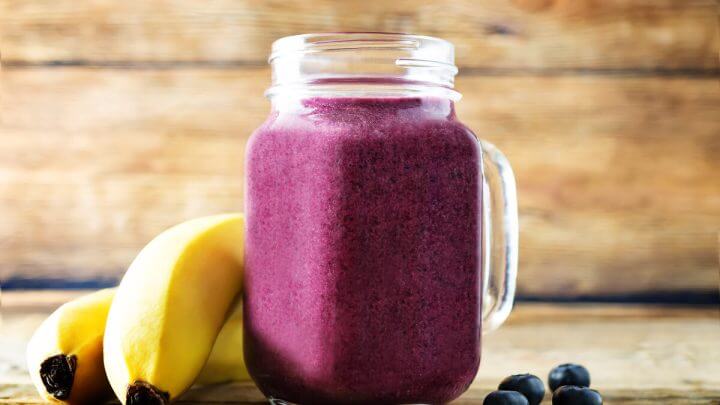 Copo de vidro com vitamina de açaí e banana em cima de uma mesa de madeira com banana e açaí ao lado