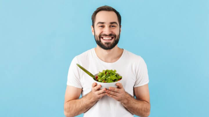 Homem vestindo blusa branca em frente a um fundo azul, sorrindo e segurando um recipiente com salada