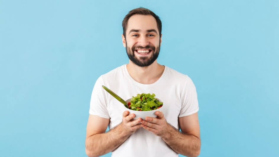 Homem vestindo blusa branca em frente a um fundo azul, sorrindo e segurando um recipiente com salada