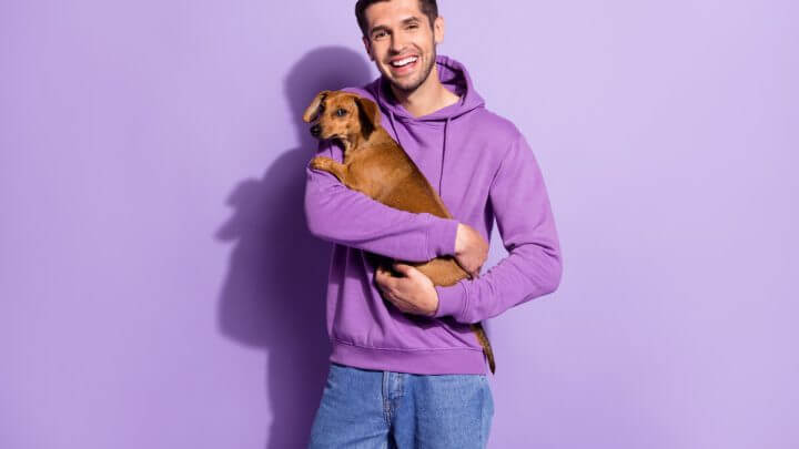 Homem vestindo blusa de moletom roxa e segurando um cachorro em frente a um fundo roxo