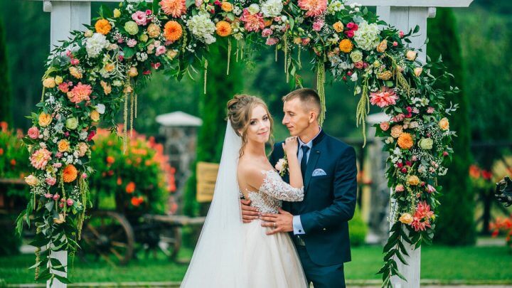 Mulher vestida de noiva e homem com terno em frente a um arco de flores abraçados