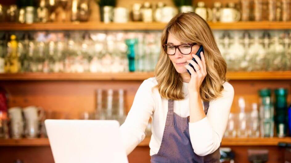 Mulher vestindo avental sentada mexendo em um computador e falando no telefone