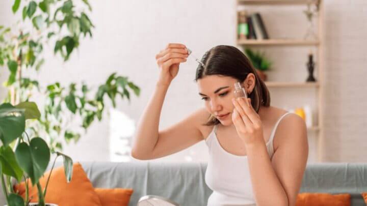 Mulher aplicando produto com conta-gotas no couro cabeludo enquanto segura potinho
