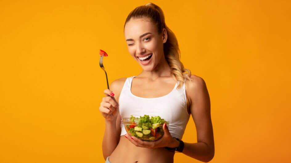 Mulher piscando e sorrindo segurando recipiente com salada e garfo com tomate