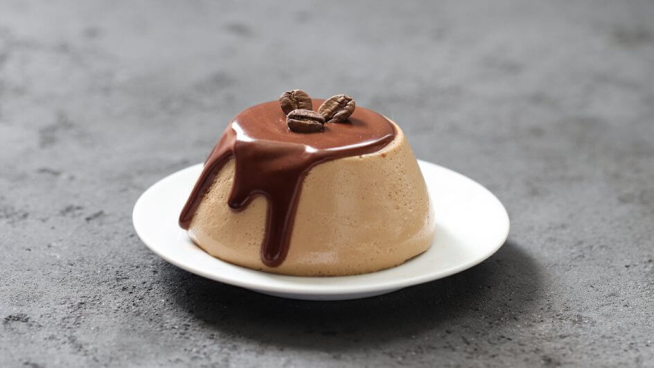 Panna cotta em cima de um prato branco decorada com calda de chocolare e grãos de café