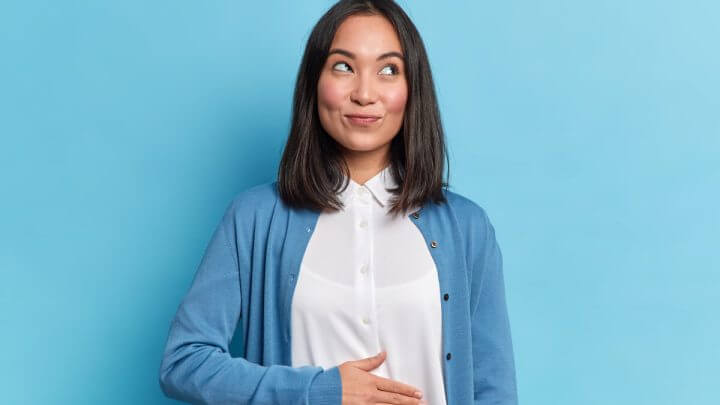 Mulher sorrindo, olhando para o lado e com a mão na barriga em frente a um fundo azul