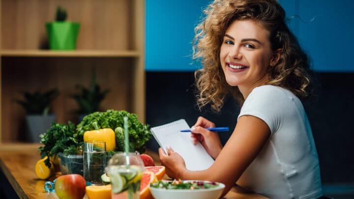 Mulher sentada com caderno e caneta na mão e alimentos saudáveis em cima da mesa