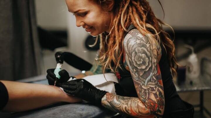 Mulher sorrindo fazendo tatuagem no braço de uma pessoa