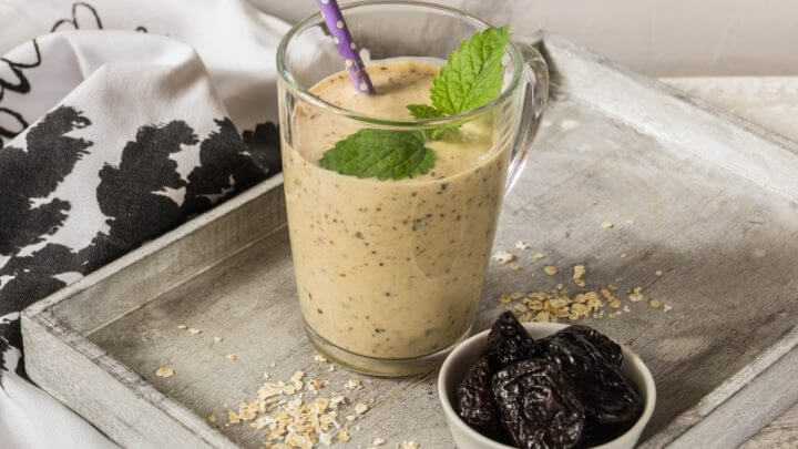 Copo de vidro com vitamina de banana e ameixa em cima de uma tábua cinza com flocos de aveia e ameixas secas