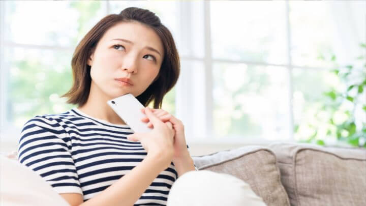 Mulher jovem sentada em um sofá e olhando para cima. Ela está segurando um celular na mão e demonstra estar esperando uma resposta.