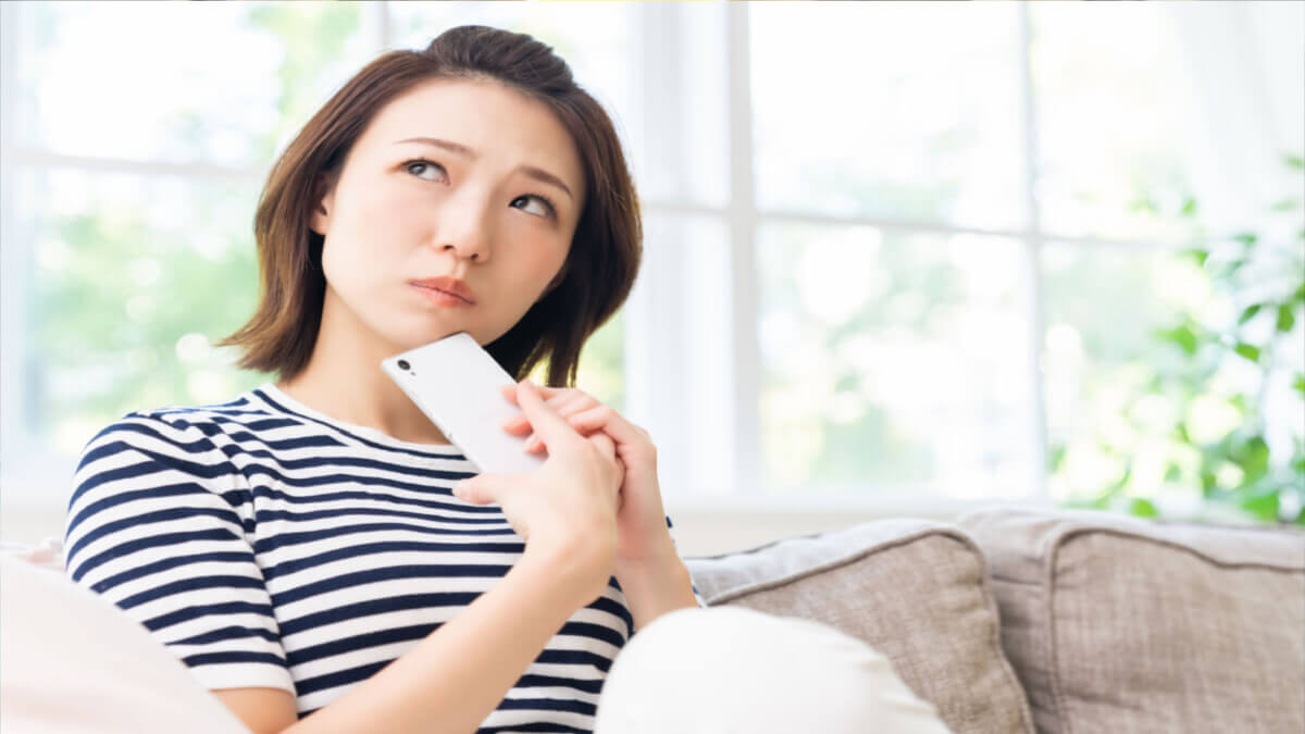Mulher jovem sentada em um sofá e olhando para cima. Ela está segurando um celular na mão e demonstra estar esperando uma resposta.