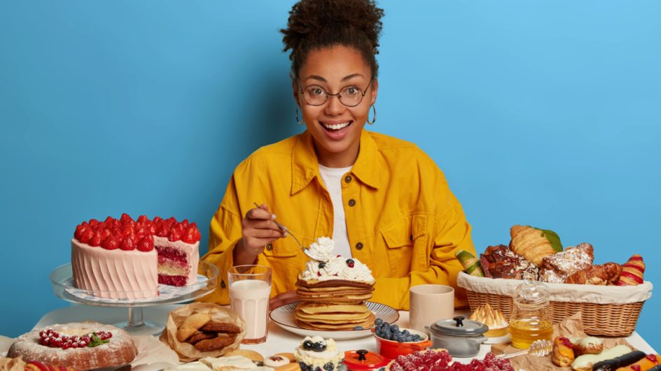 Mulher com blusa amarela de frente para uma mesa cheia de alimentos gordurosos e cheios de açúcar, tais como bolo, panqueca, croissant, tortas, chocolates e mais