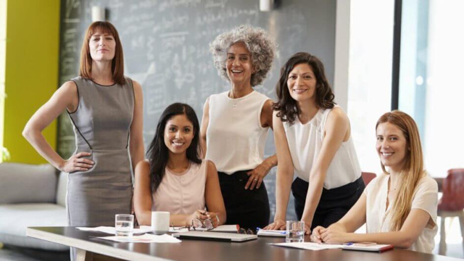 Cinco mulheres de diferentes faixas etárias sorrindo para a câmera em uma reunião de trabalho