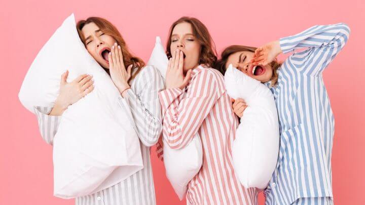 Três mulheres com pijamas, segurando travesseiros e bocejando