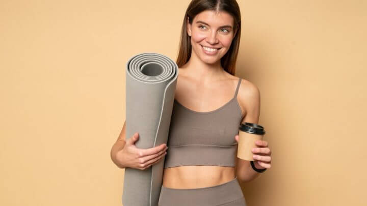 Mulher com roupa de academia sorrindo segurando um copo de café e um tapete e ioga