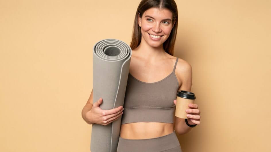 Mulher com roupa de academia sorrindo segurando um copo de café e um tapete e ioga