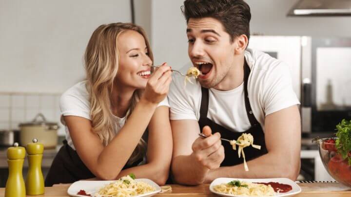 Mulher loira colocando garfo com macarrão na boca de homem. Eles estão na cozinha