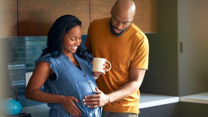 Mulher segurando caneca e com a mão na barriga. Homem ao lado dela também toca a barriga