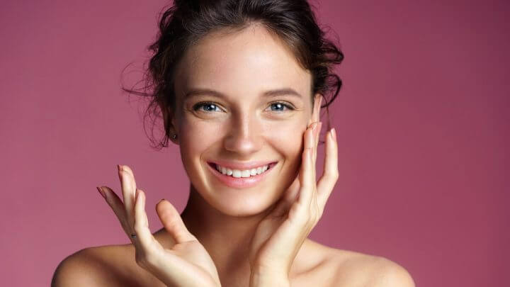 Mulher sorrindo com uma mão no rosto em frente a um fundo rosa