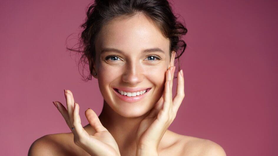 Mulher sorrindo com uma mão no rosto em frente a um fundo rosa