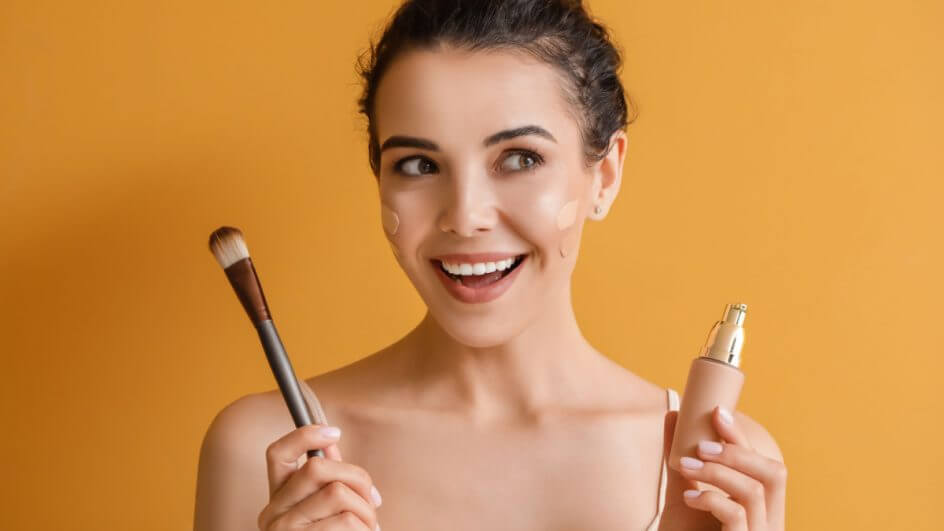 Mulher com base no rosto, segurando um pincel de maquiagem em uma mão, e uma embalagem de base na outra