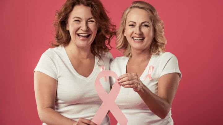 Duas mulheres sorrindo e segurando laço rosa em referência ao câncer de mama