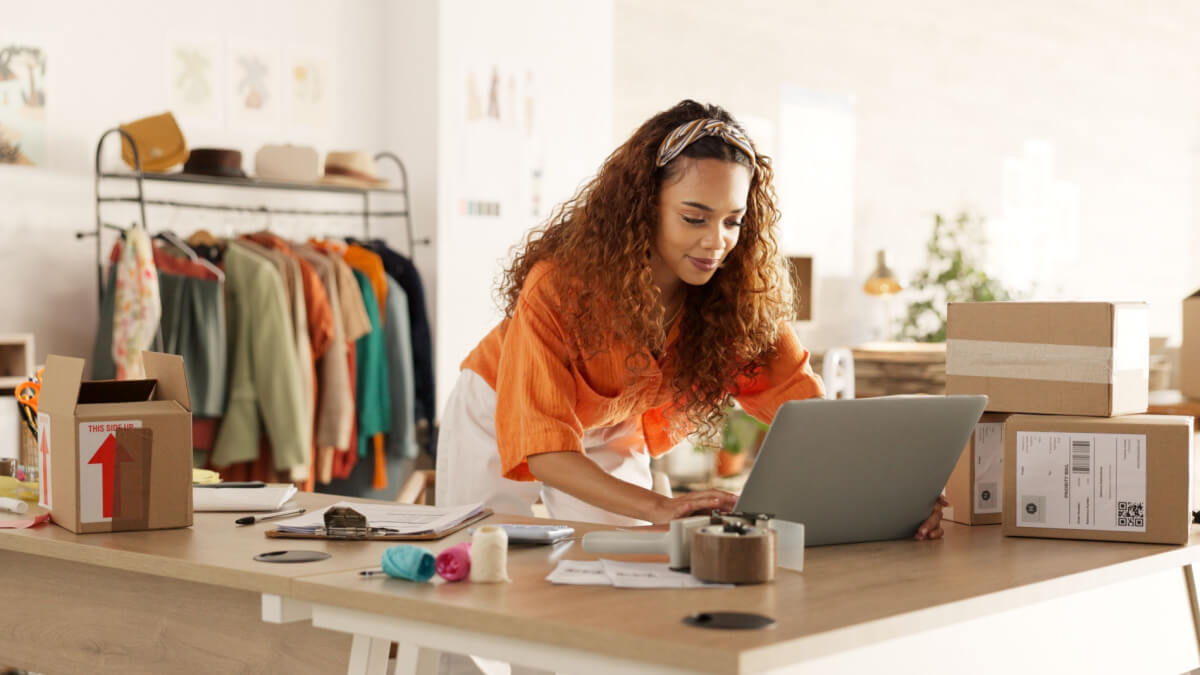Mulher negra trabalhando no laptop com caixas de encomenda ao lado e uma arara de roupas ao fundo.