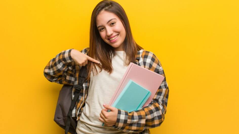 Menina sorrindo e apontando para livros em suas mãos, em um fundo amarelo