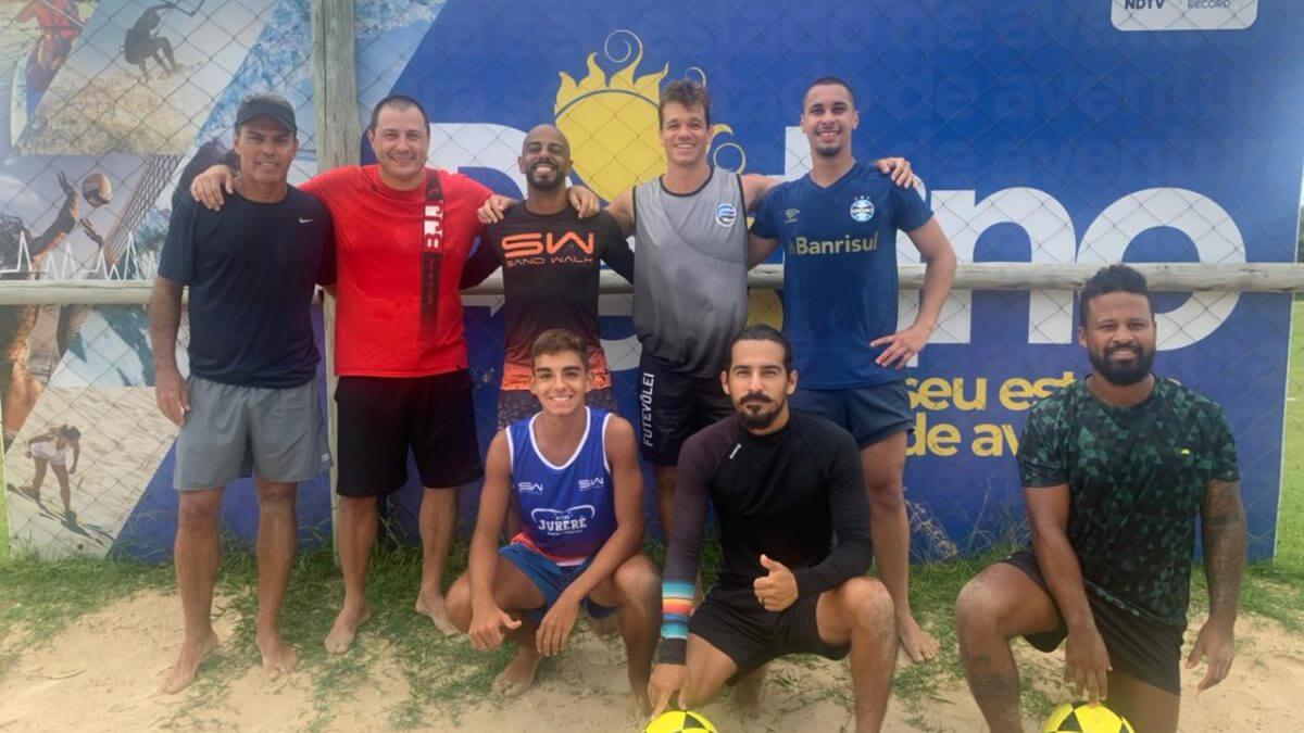 Imagem mostra cinco pessoas abraçadas em pé e outras duas agachadas e um muro azul no fundo