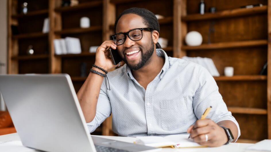 Homem negro com óculos, sorrindo e falando ao celular, enquanto olha o computador e segura um lápis com a outra mão. Ele está em uma sala com um estante marrom no fundo