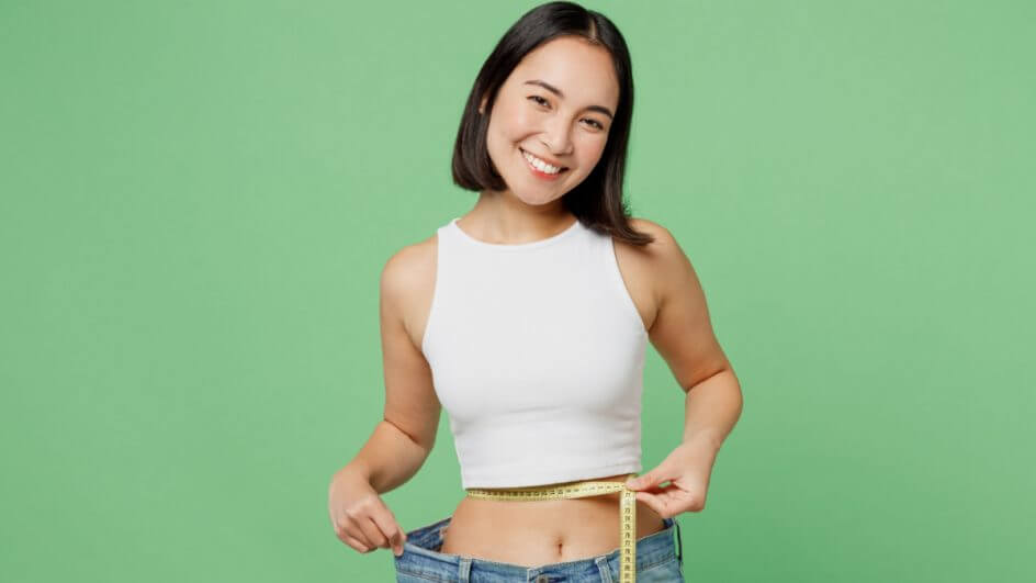 Menina se camiseta branca segurando fita métrica na barriga e calça jeans larga