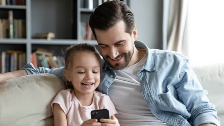 Pai e filha pequena sentados no sofá. Ela mexe em celular preto